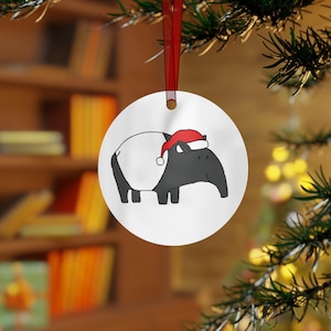 May include: A round, white Christmas ornament featuring a cartoon tapir wearing a red Santa hat. The tapir is black and white. The ornament hangs from a red ribbon against a blurred background of a Christmas tree and a bookshelf.