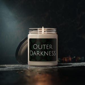 May include: A white candle in a glass jar with a black label that says "OUTER DARKNESS" in white text. The label has a green leafy pattern.