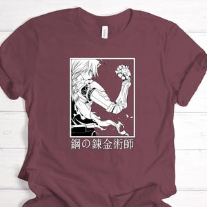 May include: Maroon t-shirt featuring a black and white illustration of an anime character with a mechanical arm. The graphic is in a white rectangle, with Japanese text below. The shirt has short sleeves and a crew neck.