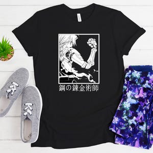 May include: Black t-shirt featuring a white and black illustration of an anime character with robotic arm and the Japanese text "鋼の錬金術師" inside a white rectangle. The shirt is paired with gray sneakers and patterned leggings.
