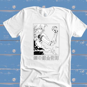 May include: White t-shirt featuring a black and white illustration of an anime character with mechanical arm and the Japanese text "鋼の錬金術師". The graphic is framed in a rectangle. The shirt is made of a soft material.