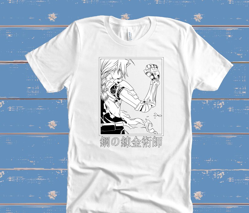 May include: White t-shirt featuring a black and white illustration of an anime character with mechanical arm and the Japanese text "鋼の錬金術師". The graphic is framed in a rectangle. The shirt is made of a soft material.