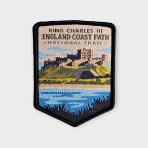 May include: A shield-shaped embroidered patch featuring a castle on a hill above a body of water. The text reads KING CHARLES III ENGLAND COAST PATH NATIONAL TRAIL. The design uses shades of blue, green, yellow, and tan with a black border.