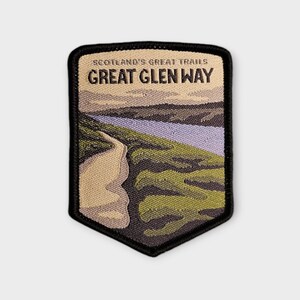 May include: A shield-shaped embroidered patch featuring a landscape of a winding path, green hills, and a light purple body of water. The text SCOTLAND'S GREAT TRAILS and GREAT GLEN WAY is stitched in black at the top against a cream-coloured sky.
