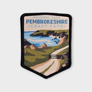 May include: A rectangular embroidered patch with a black border featuring a landscape of the Pembrokeshire Coast Path. The design includes green hills, a winding light-coloured path, blue water, and rocky cliffs with the text NATIONAL TRAIL PEMBROKESHIRE COAST PATH.