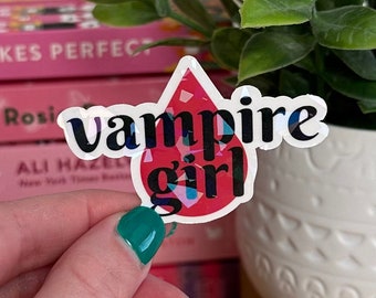 May include: A holographic sticker with a red blood drop shape. The sticker says "Vampire girl" in black text.
