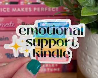 Puede incluir: Una pegatina holográfica con una pila de libros y el texto "emotional support kindle". La pegatina tiene un esquema de colores arcoíris y un diseño brillante.