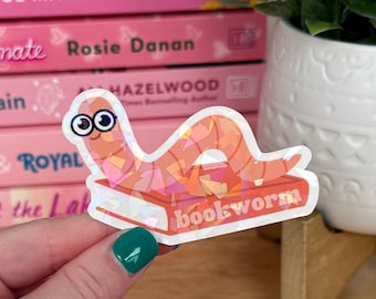 Peut inclure: Un autocollant iridescent rose et orange d'un ver sur un livre avec le texte "bookworm" sur le livre.