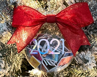 May include: A clear glass Christmas ornament with a red bow and the year 2021 printed on it. The ornament is filled with small, colorful photos.