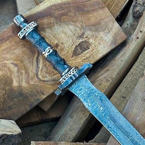 May include: A detailed image of a long, ornate sword with a textured, blue-gray blade. The handle is dark with silver-colored accents. The sword rests on a pile of various wooden planks, showcasing the craftsmanship and design of the weapon.
