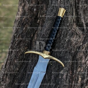 May include: A medieval-style sword with a silver blade and a gold-colored crossguard and pommel. The handle is wrapped in black material. The sword is resting against a tree trunk.
