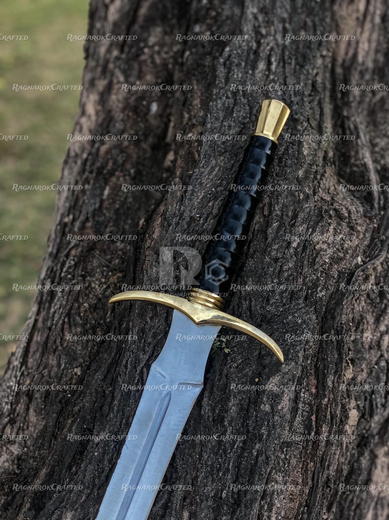 May include: A medieval-style sword with a silver blade and a gold-colored crossguard and pommel. The handle is wrapped in black material. The sword is resting against a tree trunk.