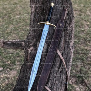 May include: A medieval-style sword with a polished, light blue blade and a gold crossguard. The handle is black with gold accents. A brown leather sheath with a strap is next to the sword. The sword is leaning against a tree trunk.