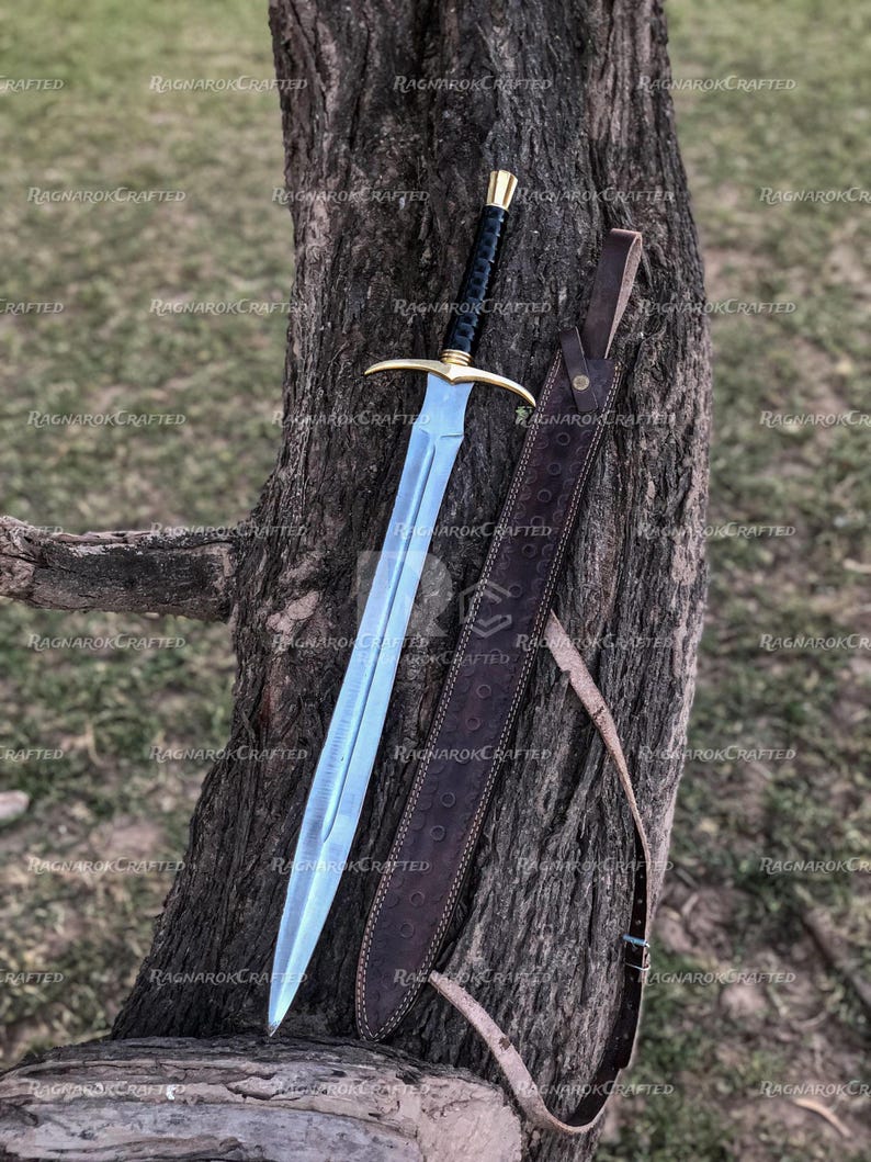 May include: A medieval-style sword with a polished, light blue blade and a gold crossguard. The handle is black with gold accents. A brown leather sheath with a strap is next to the sword. The sword is leaning against a tree trunk.