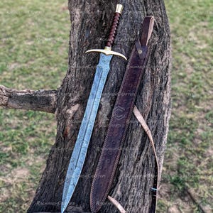 May include: A detailed image of a sword with a patterned blade and a brown leather-wrapped handle. The sword has a gold-colored crossguard and a matching pommel. A brown leather sheath with embossed detailing rests beside the sword against a tree trunk.
