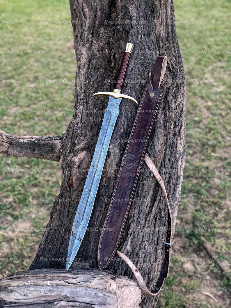 May include: A detailed image of a sword with a patterned blade and a brown leather-wrapped handle. The sword has a gold-colored crossguard and a matching pommel. A brown leather sheath with embossed detailing rests beside the sword against a tree trunk.