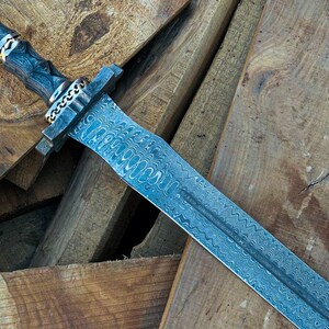 May include: A detailed image of a sword with a patterned blade and a dark wooden handle. The handle is adorned with silver and gold accents. The sword rests on a pile of wooden planks, showcasing the craftsmanship.