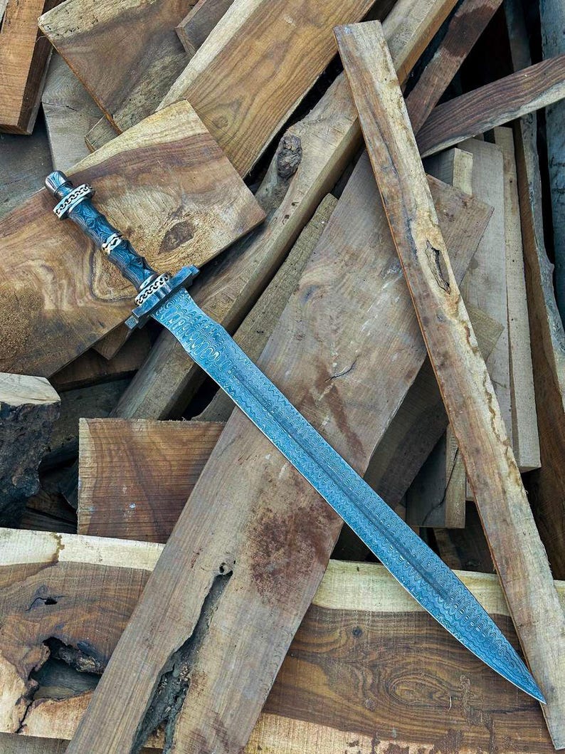 May include: A detailed image of a long, ornate sword with a textured, blue-gray blade. The handle is dark with silver-colored accents. The sword rests on a pile of various wooden planks, showcasing the craftsmanship and design of the weapon.