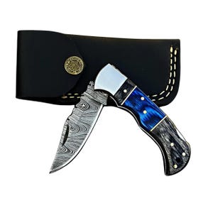 May include: A folding knife with a patterned blade and a blue and black handle. The knife is partially open, revealing the blade's design. A black leather sheath with a gold-colored button is behind the knife.