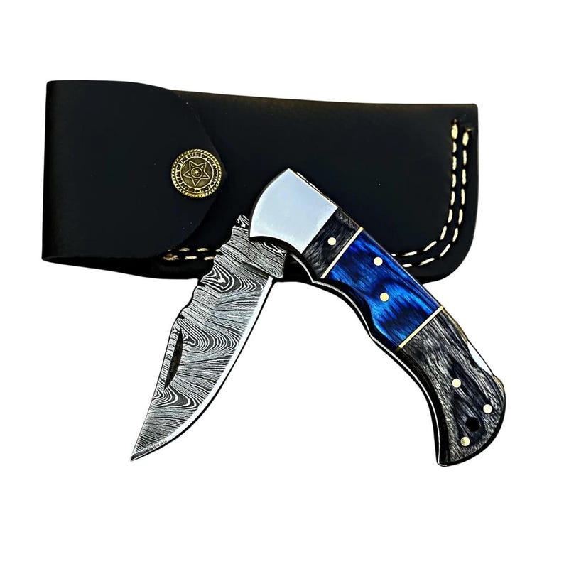 May include: A folding knife with a patterned blade and a blue and black handle. The knife is partially open, revealing the blade's design. A black leather sheath with a gold-colored button is behind the knife.