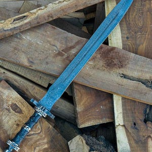 May include: A long, ornate sword with a blue-toned blade and a detailed, patterned design. The hilt is dark with silver and gold accents. The sword rests on a pile of rough-hewn wooden planks.