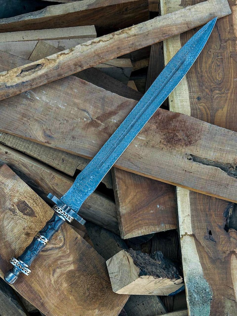 May include: A long, ornate sword with a blue-toned blade and a detailed, patterned design. The hilt is dark with silver and gold accents. The sword rests on a pile of rough-hewn wooden planks.