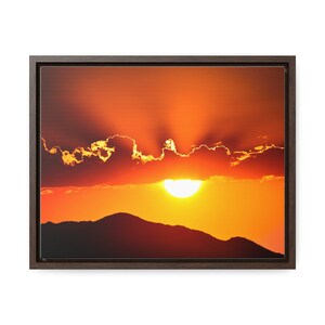 Puede incluir: Una impresión en lienzo enmarcada de una puesta de sol sobre una cordillera. El cielo es de un naranja y rojo vibrantes con un sol amarillo brillante que brilla a través de las nubes.