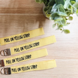 May include: Four yellow keychains with the text "PULL ON YELLOW STRAP" in black. Each keychain has a silver-colored metal clasp and ring. The keychains are arranged on a light wood surface, with a small green plant in the background.