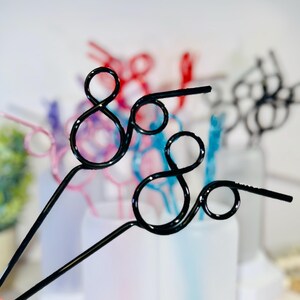 May include: Black metal decorative straws with a unique looped design. The straws are in focus, with other colorful straws in the background. The straws are a fun and stylish alternative to traditional straws.