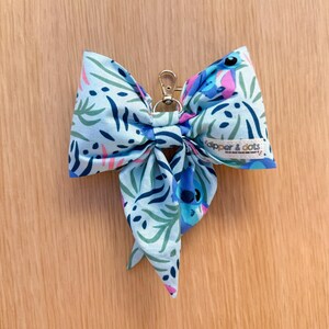 May include: A light blue fabric bow with a gold-colored clasp. The bow features a pattern of green, blue, and pink botanical elements. The tag reads "dipper & dots".