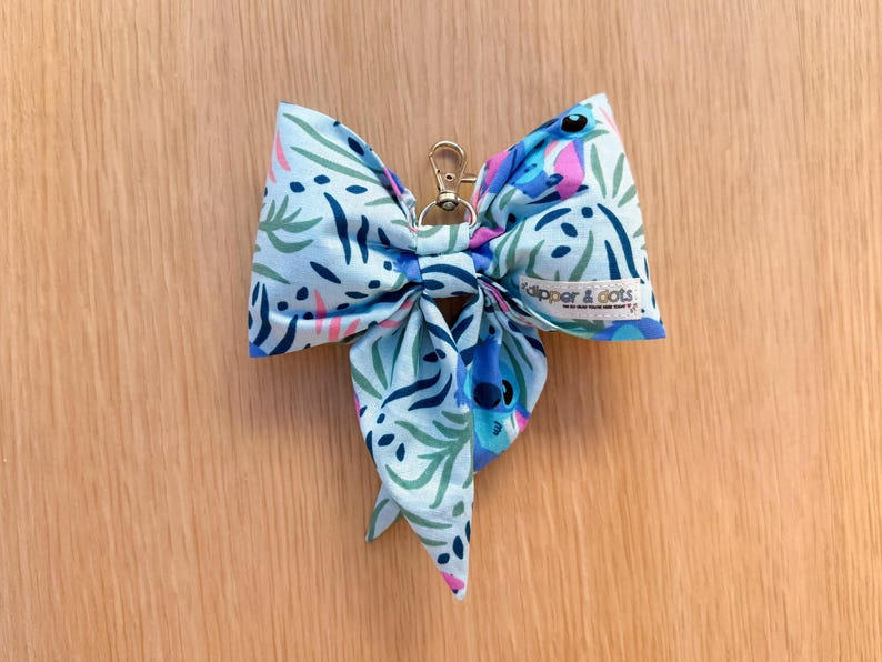 May include: A light blue fabric bow with a gold-colored clasp. The bow features a pattern of green, blue, and pink botanical elements. The tag reads "dipper & dots".