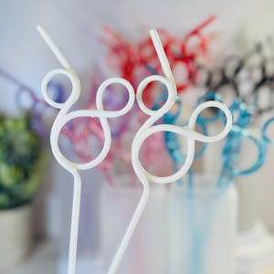 May include: White, swirly plastic straws with a decorative design. Other straws in the background are red, pink, blue, and black. The straws are in a clear container, with a small green plant in a pot to the left.