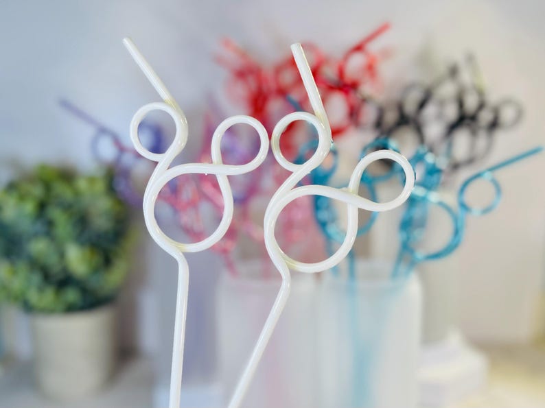 May include: White, swirly plastic straws with a decorative design. Other straws in the background are red, pink, blue, and black. The straws are in a clear container, with a small green plant in a pot to the left.