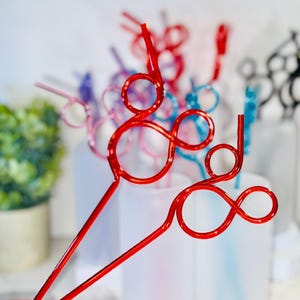 May include: A collection of colorful, decorative straws in various shades, including red, pink, blue, and black. The straws have a unique looped design, perfect for adding a festive touch to beverages. The background is blurred, highlighting the straws.