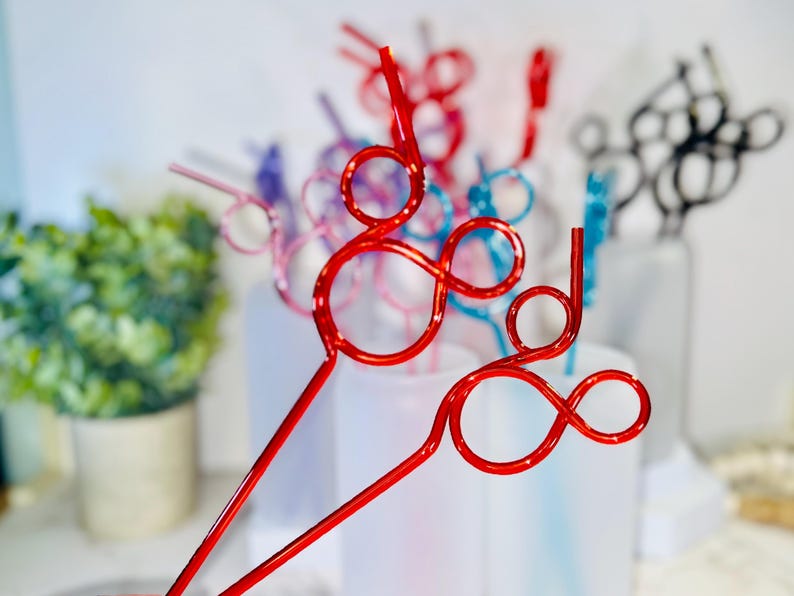 May include: A collection of colorful, decorative straws in various shades, including red, pink, blue, and black. The straws have a unique looped design, perfect for adding a festive touch to beverages. The background is blurred, highlighting the straws.