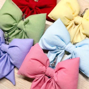 May include: A collection of colorful fabric bows in various shades, including red, green, yellow, blue, pink, and purple. Each bow is tied and features a silver-colored metal clip. The bows are arranged on a light-colored wooden surface.