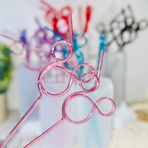 May include: Pink metal drink stirrers with a looped design. Other colorful stirrers in red, blue, and black are visible in the background. The stirrers are designed for beverages and add a decorative touch.