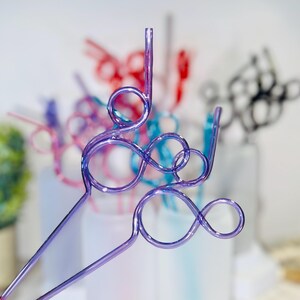 May include: Close-up of several colorful, decorative drinking straws. The straws are made of clear plastic and feature a looped design. The straws are in shades of purple, red, pink, blue, and black. The background is blurred, with a hint of greenery.