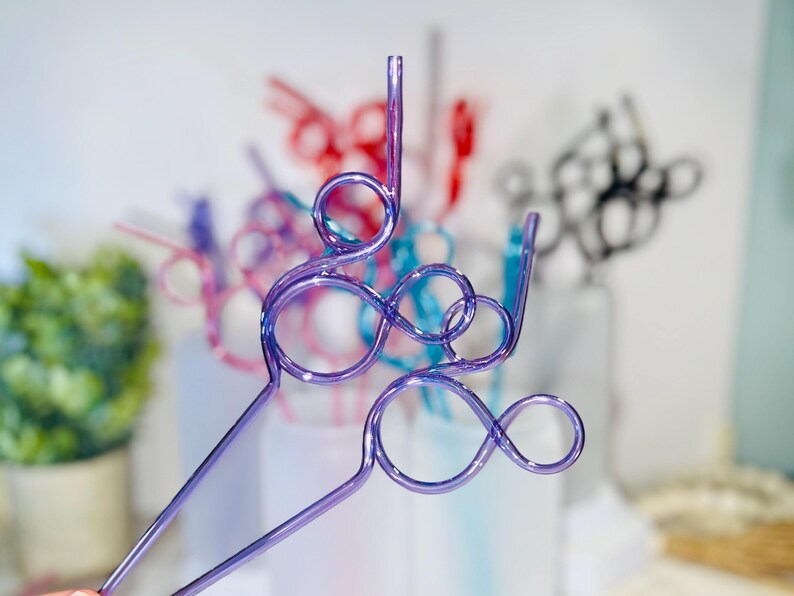 May include: Close-up of several colorful, decorative drinking straws. The straws are made of clear plastic and feature a looped design. The straws are in shades of purple, red, pink, blue, and black. The background is blurred, with a hint of greenery.