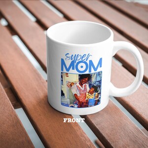 以下が含まれることがあります： 白いセラミック製のマグカップで、青と白のデザインで「スーパーママ」と書かれており、母親と子供の画像が描かれています。