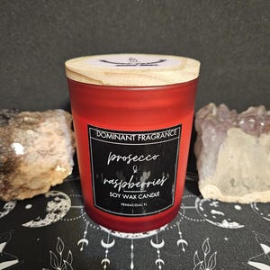 May include: Red soy wax candle with a wooden lid. The label reads "Dominant Fragrance Prosecco & Raspberries Soy Wax Candle Pensacola, FL."