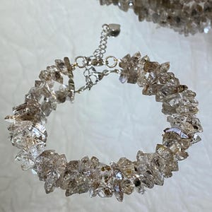 Puede incluir: Una pulsera compuesta por numerosos fragmentos pequeños de cristal en bruto, translúcidos, de color marrón y transparente. La pieza cuenta con una cadena extensora de metal plateado con un pequeño dije en forma de corazón y un cierre de mosquetón.