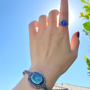 May include: A silver-toned ring and bracelet set. The ring features a round, blue stone set in an ornate, filigree design. The bracelet has a similar design with a larger, oval blue stone. The jewelry is displayed against a bright blue sky.