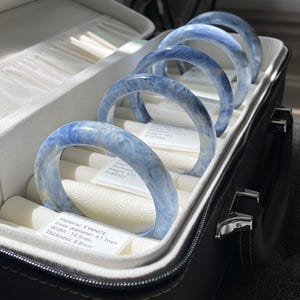 May include: A collection of blue kyanite bracelets displayed in a black jewelry case. The bracelets have an inner diameter of 61.1mm, a width of 14.5mm, and a thickness of 8.8mm. The case is open, revealing the bracelets resting on a white interior.