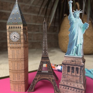 Op de afbeelding: Papieren modellen van bekende bezienswaardigheden: Big Ben, de Eiffeltoren en het Vrijheidsbeeld. Het Big Ben-model is bruin met een klok. De Eiffeltoren is bruin en het Vrijheidsbeeld is lichtblauw.