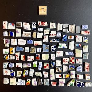 May include: A collection of broken ceramic pieces with various floral and geometric patterns, arranged on a black surface. A wooden tile with the letter "T" and the number "1" is in the upper right corner. The fragments display colors like red, blue, and white.