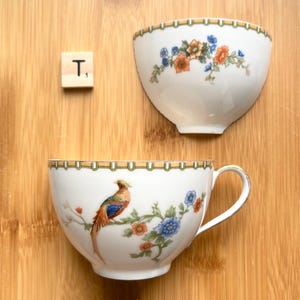May include: Two white teacups with floral and bird designs. One cup has a handle, the other is a half-cup. Both have a gold rim and are decorated with orange, blue, and green flowers and a pheasant. A wooden tile with the letter "T" is also present.