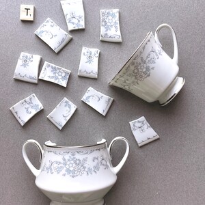 May include: White teacup and sugar bowl with a floral design in blue and gray, with silver trim. Several broken pieces of the teacup are scattered around the intact pieces. A wooden tile with the letter "T" is also present.