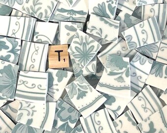 May include: A collection of white ceramic mosaic tiles featuring a light blue floral and striped pattern, interspersed with a single square wooden tile marked with the letter T and a small number one in the bottom right corner.