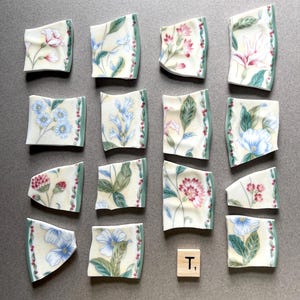 May include: Sixteen pieces of broken porcelain with floral patterns in pink, blue, and white, with green and red accents. The pieces are arranged on a gray surface. A wooden tile with the letter "T" and the number "1" is also present.